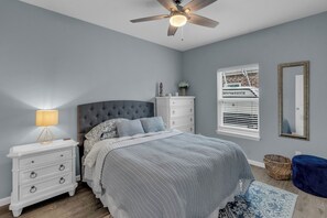 Ein Schlafzimmer mit einem Bett, Nachttischen, einer Kommode, einem Deckenventilator, einem Fenster mit Jalousien, einem Spiegel und einem blauen Hocker.