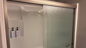 Casa de banho com chuveiro, porta de vidro e duas garrafas de shampoo numa prateleira.