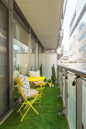 Een balkon met kunstgras, geel meubilair en potplanten.
