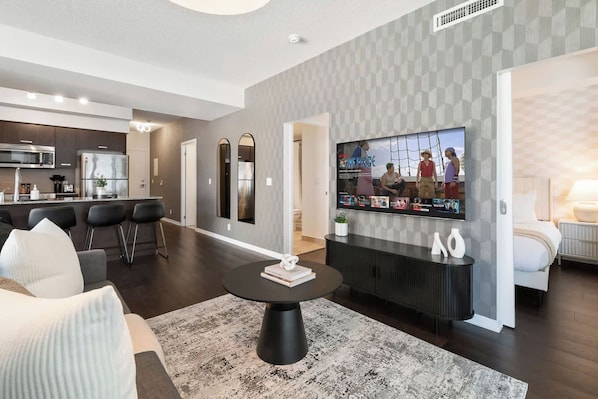 A modern living room with a round coffee table, a flat-screen TV mounted on the wall, and a kitchen in the background.