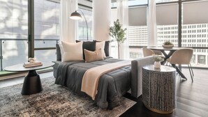 Une chambre d’hôtel moderne avec un grand lit, une table d’appoint ronde, une petite table ronde et une vue sur la ville grâce à de grandes fenêtres.