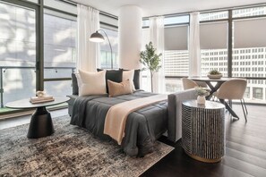 A modern hotel room with a large bed, a round side table, a small round table, and a view of the city through large windows.