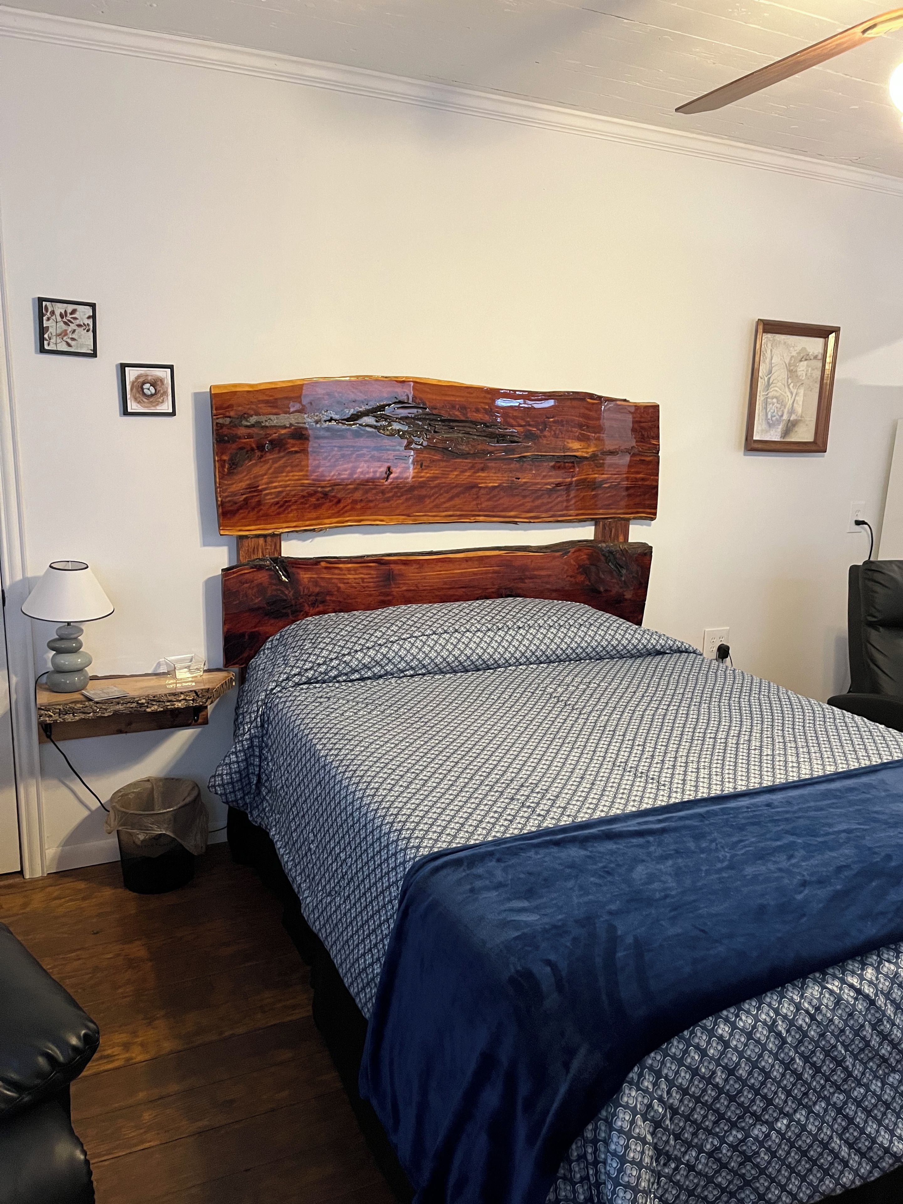 A bedroom with a wooden headboard, a bedside table with a lamp, a trash bin, and a chair.