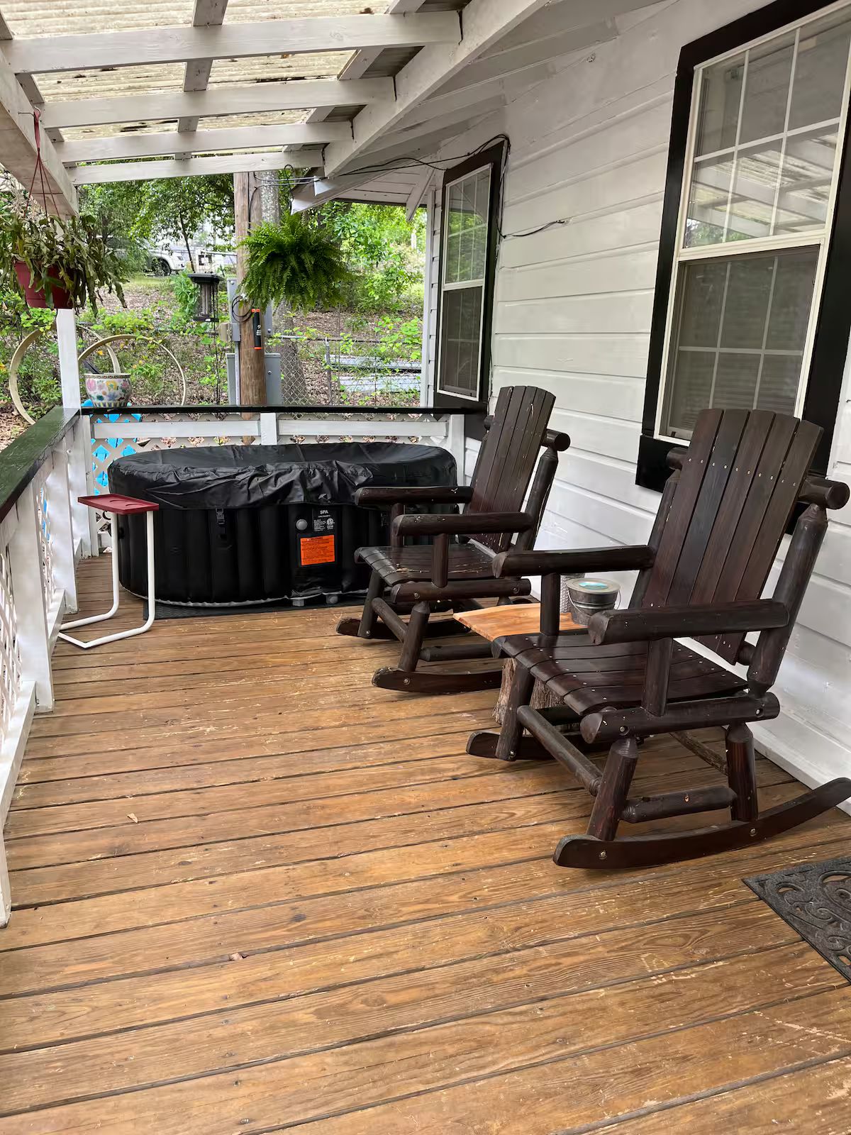 Een overdekt terras met houten vloer, twee schommelstoelen, een barbecue en een hangplanter.