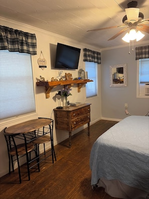 A bedroom with a bed, a desk with a chair, a television, a ceiling fan, and a window with blinds.