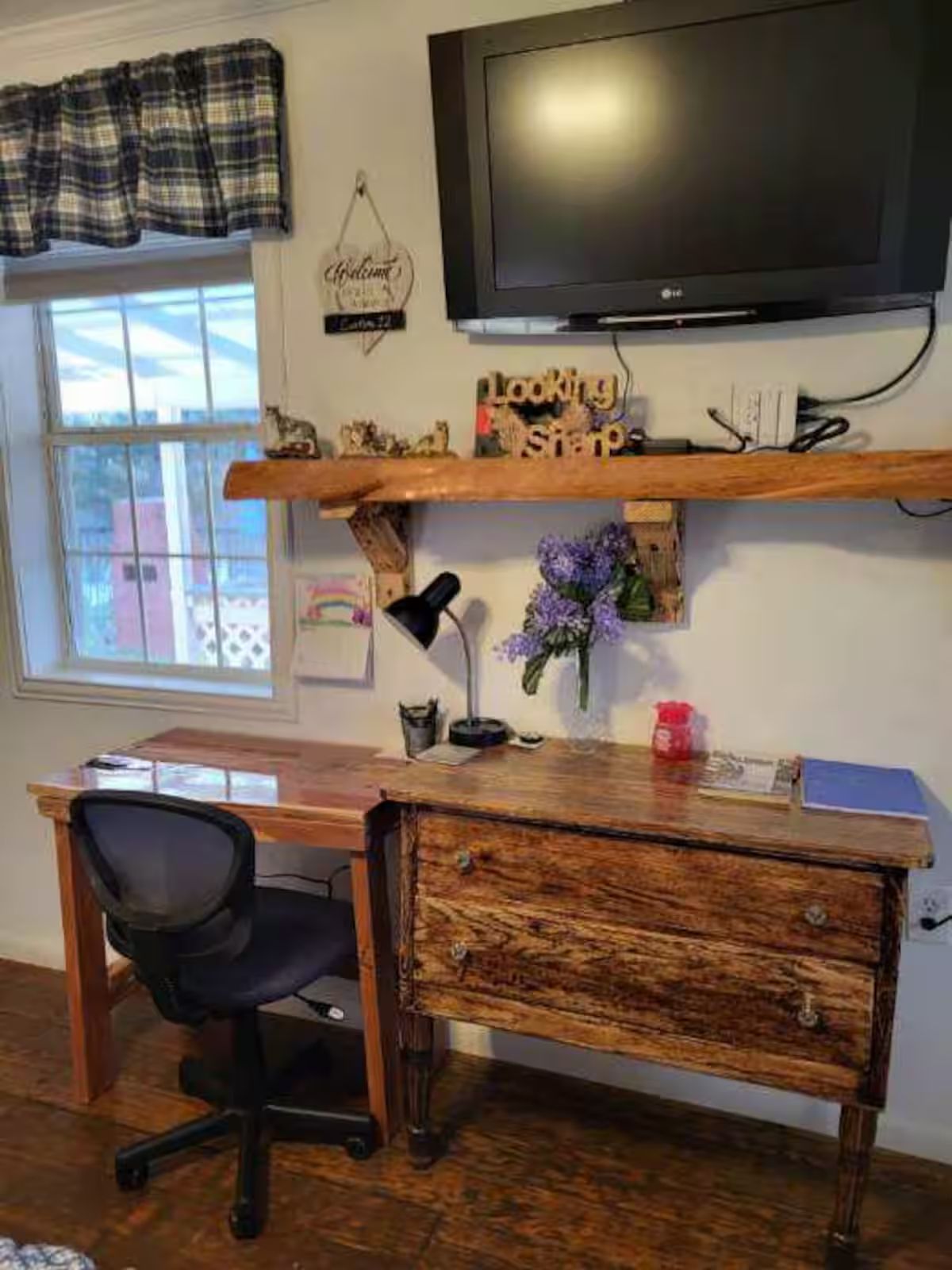 A home office with a wooden desk, a black office chair, a television mounted above the desk, and a window with a view of buildings.