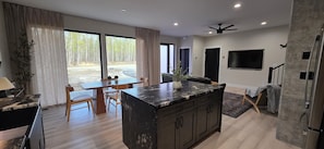 A modern kitchen with a large island, dining area with wooden chairs, and a view of a forest through large windows.