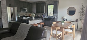 A modern kitchen with dark cabinets, a stainless steel refrigerator, and a microwave. There is a dining area with a wooden table and chairs, and a large window providing a view of trees.