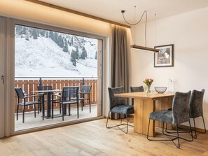 Ein Essbereich mit einem Holztisch und Stühlen, Blick auf einen verschneiten Berg durch große Glasfenster, und ein gerahmtes Bild an der Wand.