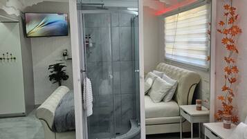 A modern bathroom with a glass shower enclosure, a white vanity, and a flat-screen TV mounted on the wall.