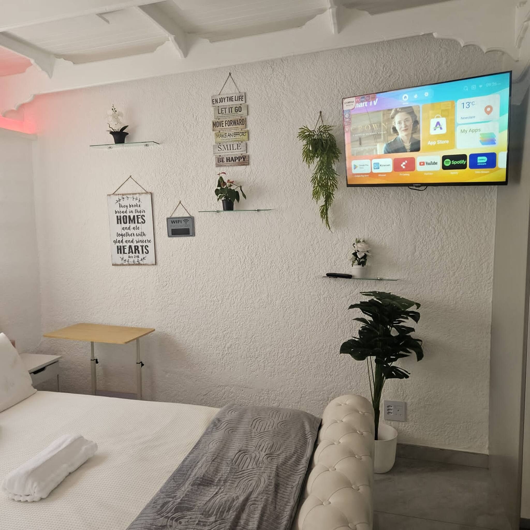 A modern bedroom with a bed, a small table, a TV mounted on the wall, and various wall decorations including a sign and a potted plant.