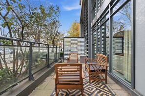 Een balkon met houten stoelen en een kamerkleed, met uitzicht op een straat met bomen en gebouwen.