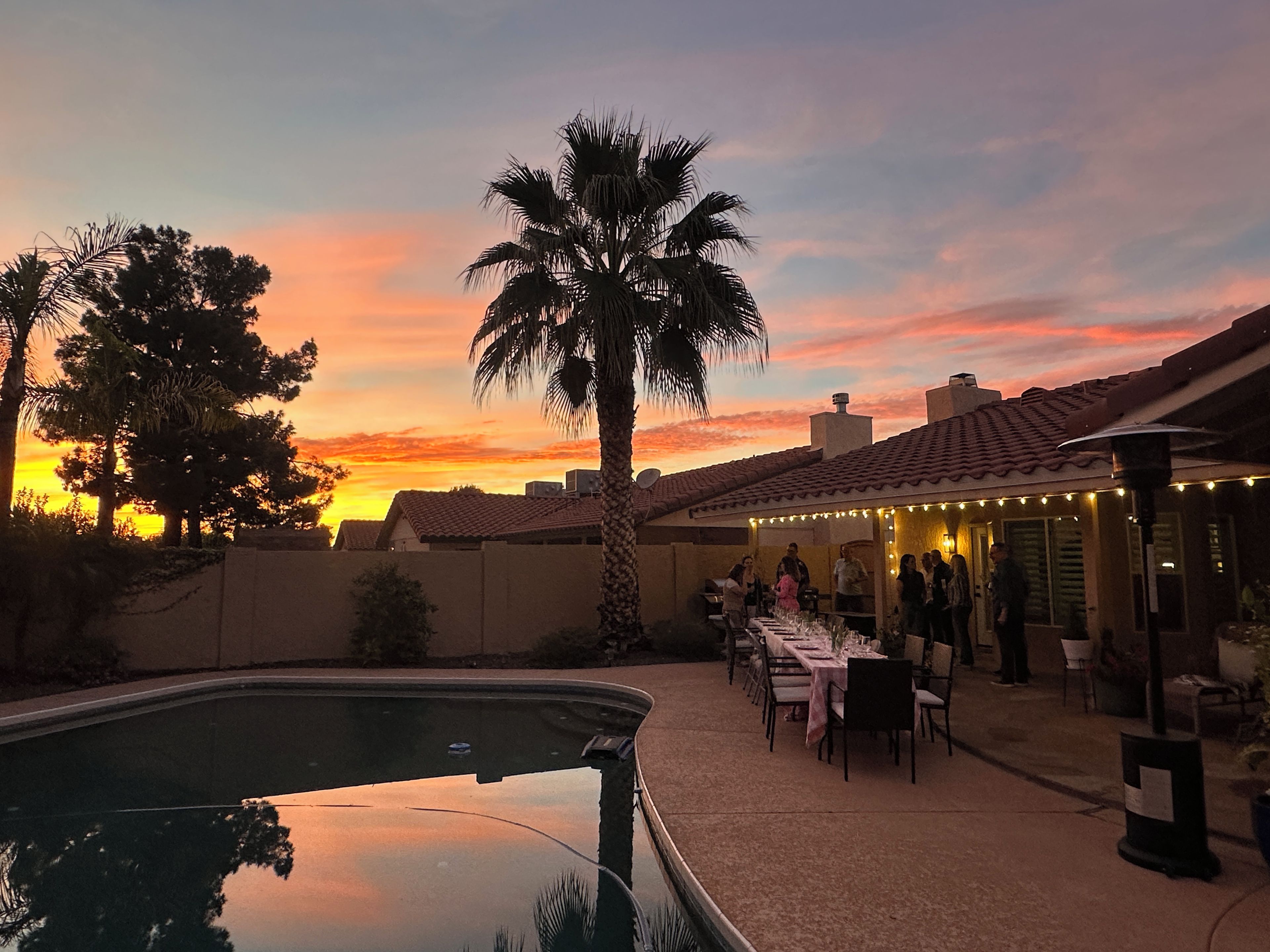 Zona junto a la piscina con vista al atardecer, mobiliario para comer al aire libre y una palmera.