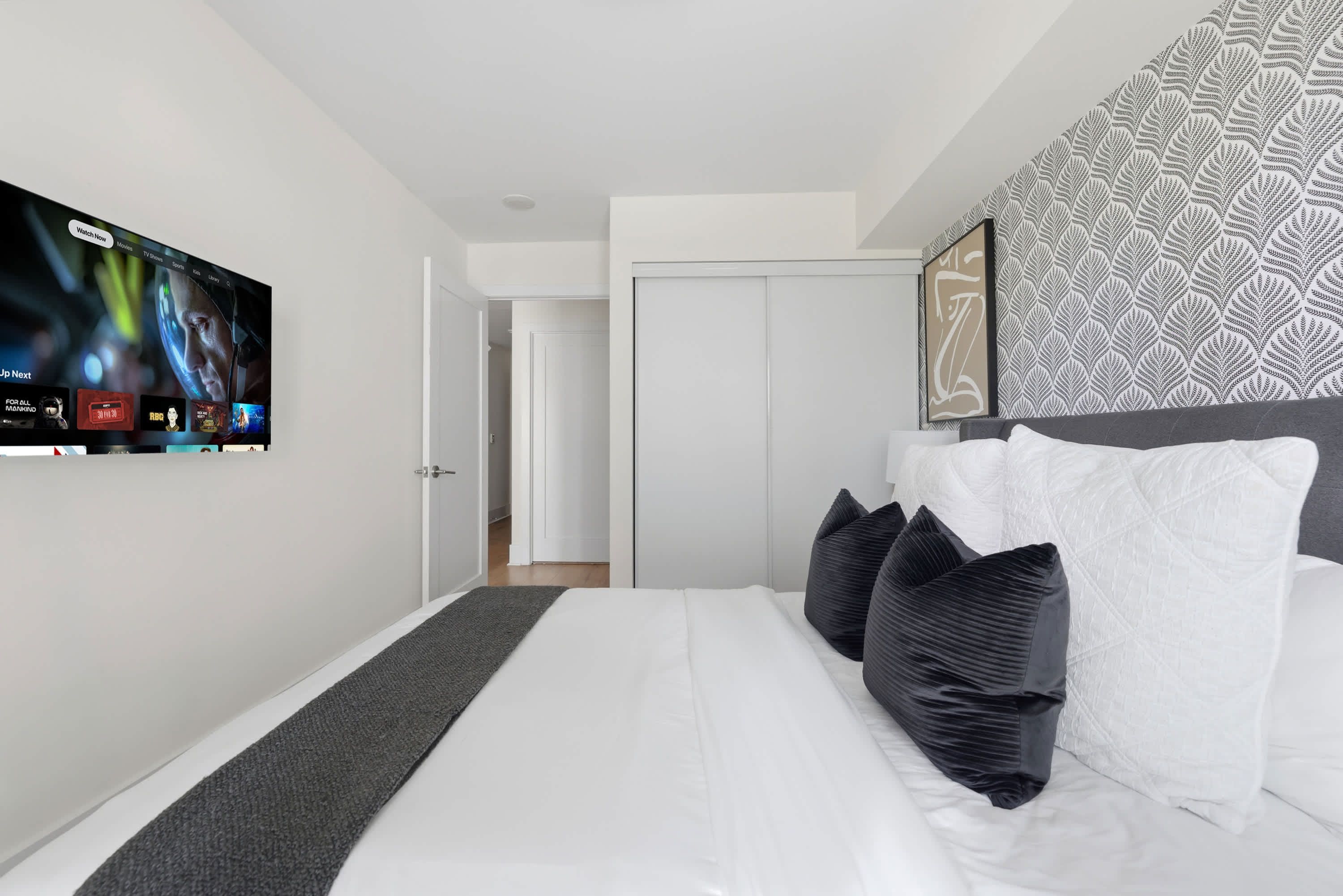 A modern hotel room with a large flat-screen TV mounted on the wall, a neatly made bed with white and dark pillows, a patterned headboard, and a door leading to a hallway.