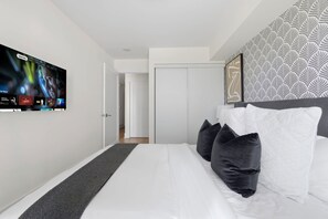 Quarto de hotel moderno com uma grande TV de tela plana na parede, uma cama bem arrumada com travesseiros brancos e escuros, cabeceira estampada e uma porta que dá para um corredor.