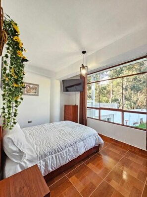 A bedroom with a bed, wooden flooring, a window with a view of trees, and a hanging plant. - Hotel Catañuñi  (Sapallanga)