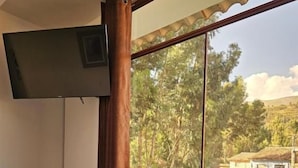 A bedroom with a bed, a window with a view of a green lawn and trees, and a mounted television. - Hotel Catañuñi  (Huancayo)