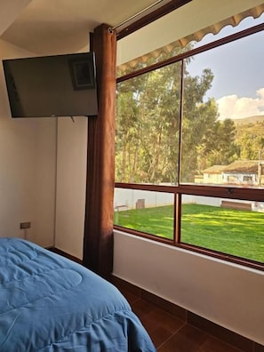 A bedroom with a bed, a window with a view of a green lawn and trees, and a mounted television. - Hotel Catañuñi  (Huancayo)