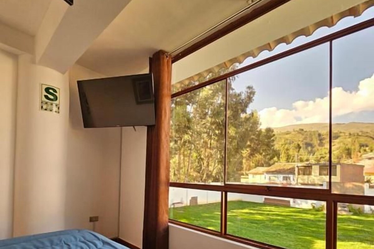 A bedroom with a bed, a television on the wall, and a large window with a view of a green landscape.