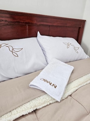 A bed with a white pillow featuring a leaf design, a white towel with 'CATANUNI' text, and a beige blanket with a white trim. - Hotel Catañuñi  (Huancayo)