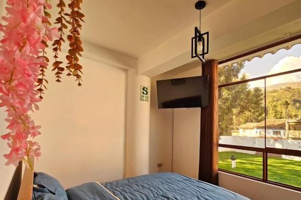 A bedroom with a bed, a TV, and a window with a view of greenery.