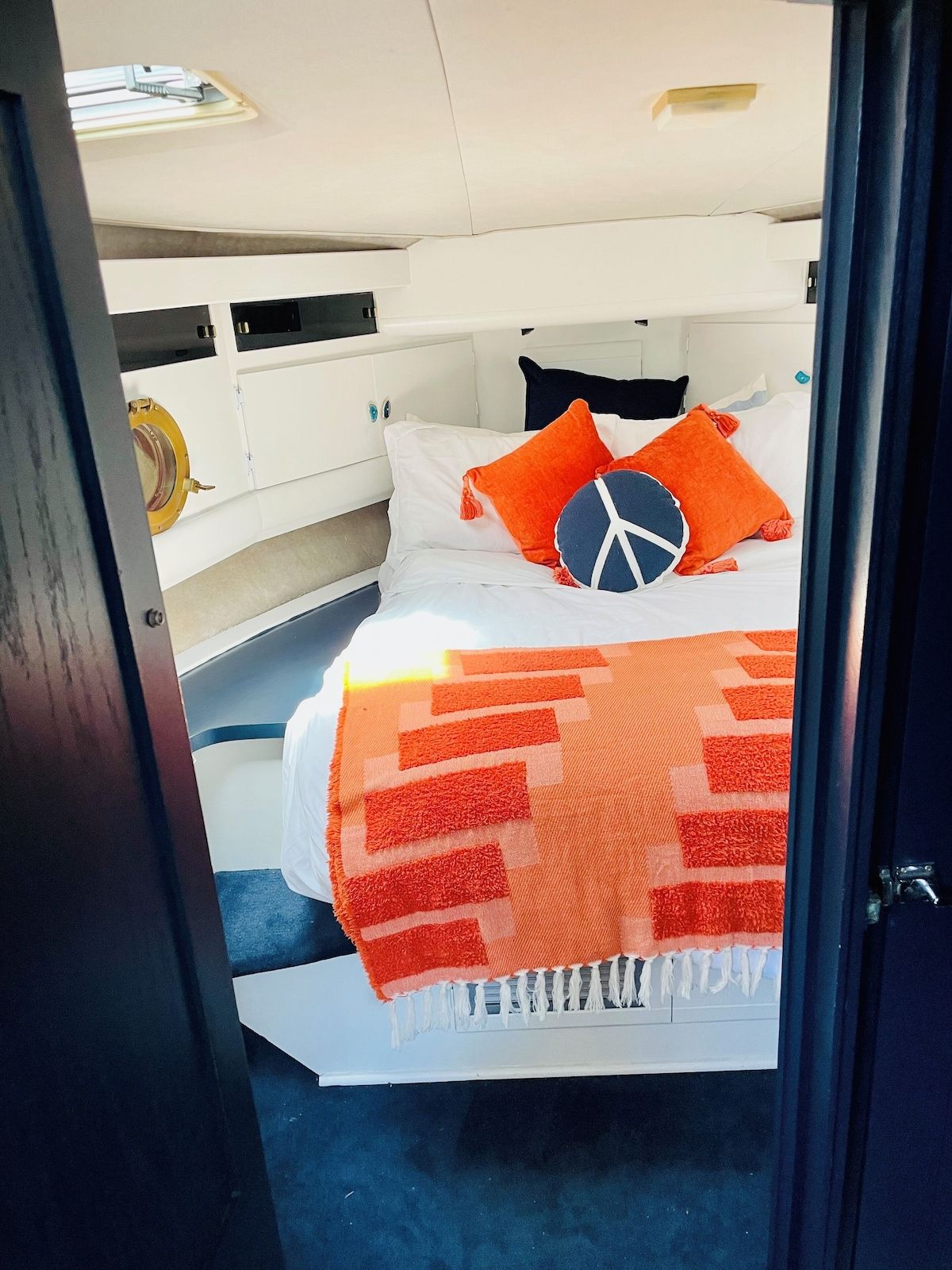 A bed with white bedding, orange pillows, and a blue peace sign pillow. There is a white headboard and a window above the bed.