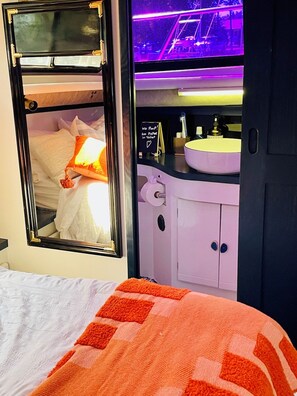 Un dormitorio con cama, ventana, espejo, mesita de noche con lámpara y baño con lavamanos e inodoro.