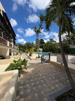 A pool area with lounge chairs, palm trees, and a building with balconies. - New Condo near Brownes Beach – Pool, Gym & Parking (Bridgetown)