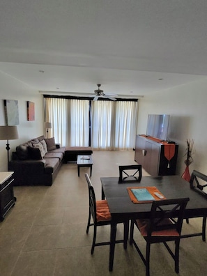 A living room with a sofa, a dining table with chairs, a TV, and a ceiling fan. - New Condo near Brownes Beach – Pool, Gym & Parking (Bridgetown)