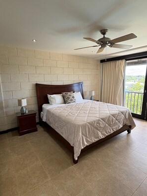 A bedroom with a bed, nightstand, ceiling fan, and a view through a window. - New Condo near Brownes Beach – Pool, Gym & Parking (Bridgetown)