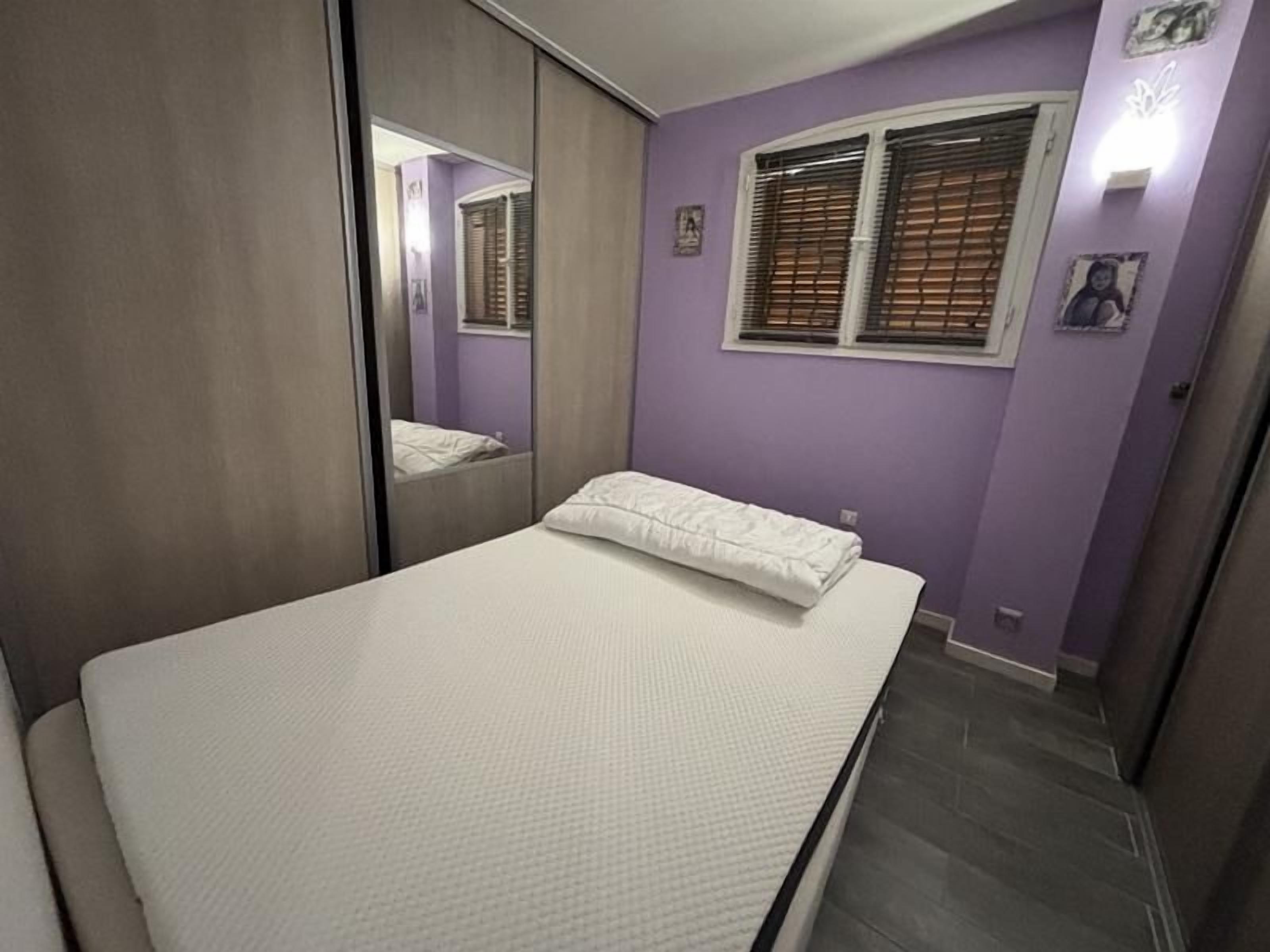 A bedroom with a bed, a window with blinds, a mirror, and a wall-mounted light fixture.