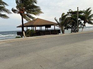 A coastal road with a wooden gazebo, palm trees, and the ocean in the background. - 2-bedroom apartment in the heart of Pensacola Roatan with cool ocean breeze (Roatan)