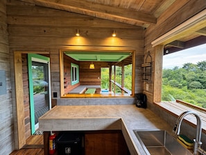 A wooden cabin interior with a kitchen area, a window with a view of greenery, and a door leading outside.