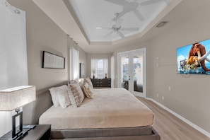 Une chambre à coucher avec un lit, un bureau avec un ordinateur, un ventilateur de plafond et un téléviseur fixé au mur.