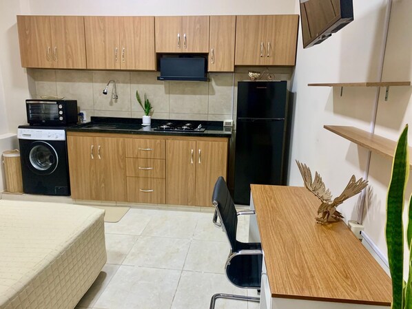 Cocina compacta con armarios de madera, microondas, lavadora, refrigerador y comedor con mesa y sillas.