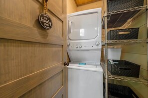 A laundry room with a white washing machine and dryer, a wooden door, and shelves with baskets. - Lux Cabin, Hot Tub, Arcade, In and Out Fireplaces (Sevierville)