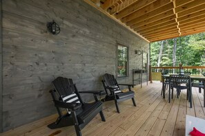A covered wooden deck with Adirondack chairs, a dining table, and a view of the surrounding forest. - Lux Cabin, Hot Tub, Arcade, In and Out Fireplaces (Sevierville)