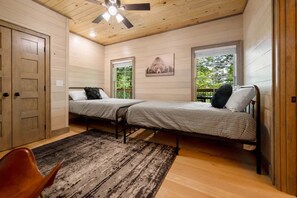 A bedroom with two beds, a ceiling fan, a wooden door, and a window with a view of greenery. - Lux Cabin, Hot Tub, Arcade, In and Out Fireplaces (Sevierville)