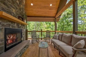 A covered outdoor seating area with a stone fireplace, wooden furniture, and a view of the surrounding trees. - Lux Cabin, Hot Tub, Arcade, In and Out Fireplaces (Sevierville)