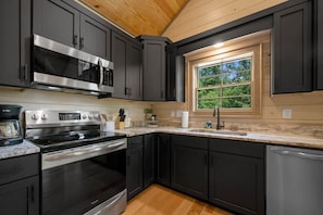 A modern kitchen with dark cabinetry, stainless steel appliances, and a window providing natural light. - Lux Cabin, Hot Tub, Arcade, In and Out Fireplaces (Sevierville)