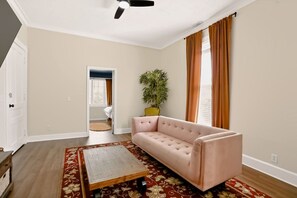 Une pièce avec un canapé beige, une table basse en bois, une plante en pot et une vue sur l’extérieur par une porte ouverte.