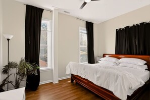 A bedroom with a wooden bed, two windows with black curtains, a ceiling fan, and a potted plant. - Spacious Herron-Morton Stay Near Downtown (Indianapolis)