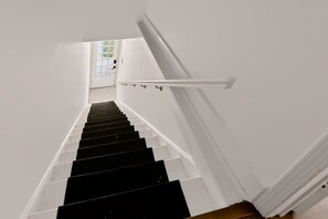 Eine Treppe mit weißen Wänden und schwarzen Stufen, die zu einer Tür mit Blick auf Grünanlagen führt.