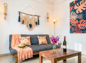 Ein modernes Wohnzimmer mit einem grauen Sofa, floralen Kissen, einem Holztisch, einer Vase mit violetten Blumen, einer Weinflasche und einer Stehlampe.