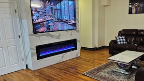 Een woonkamer met een flatscreen televisie aan een witte muur, een haard eronder en een leren bank met een sierkussen.