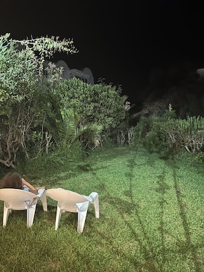 Un espace extérieur aménagé pour la nuit, avec une pelouse verdoyante, une personne assise sur une chaise, ainsi que diverses plantes et arbustes.