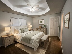 Ein Schlafzimmer mit einem Bett, Nachttischen, einer Stehlampe, einem Deckenventilator, einer Tür und einer gerahmten Pflanze an der Wand.