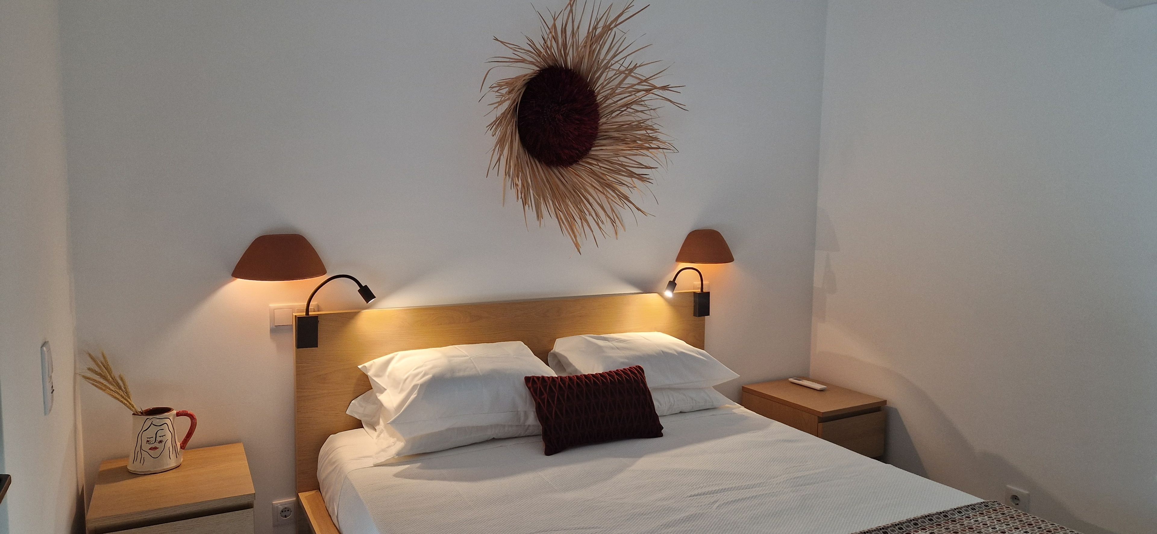 Un lit bien fait, avec du linge de lit blanc et un seul coussin décoratif, flanqué de deux tables de chevet sur lesquelles sont posées des lampes.