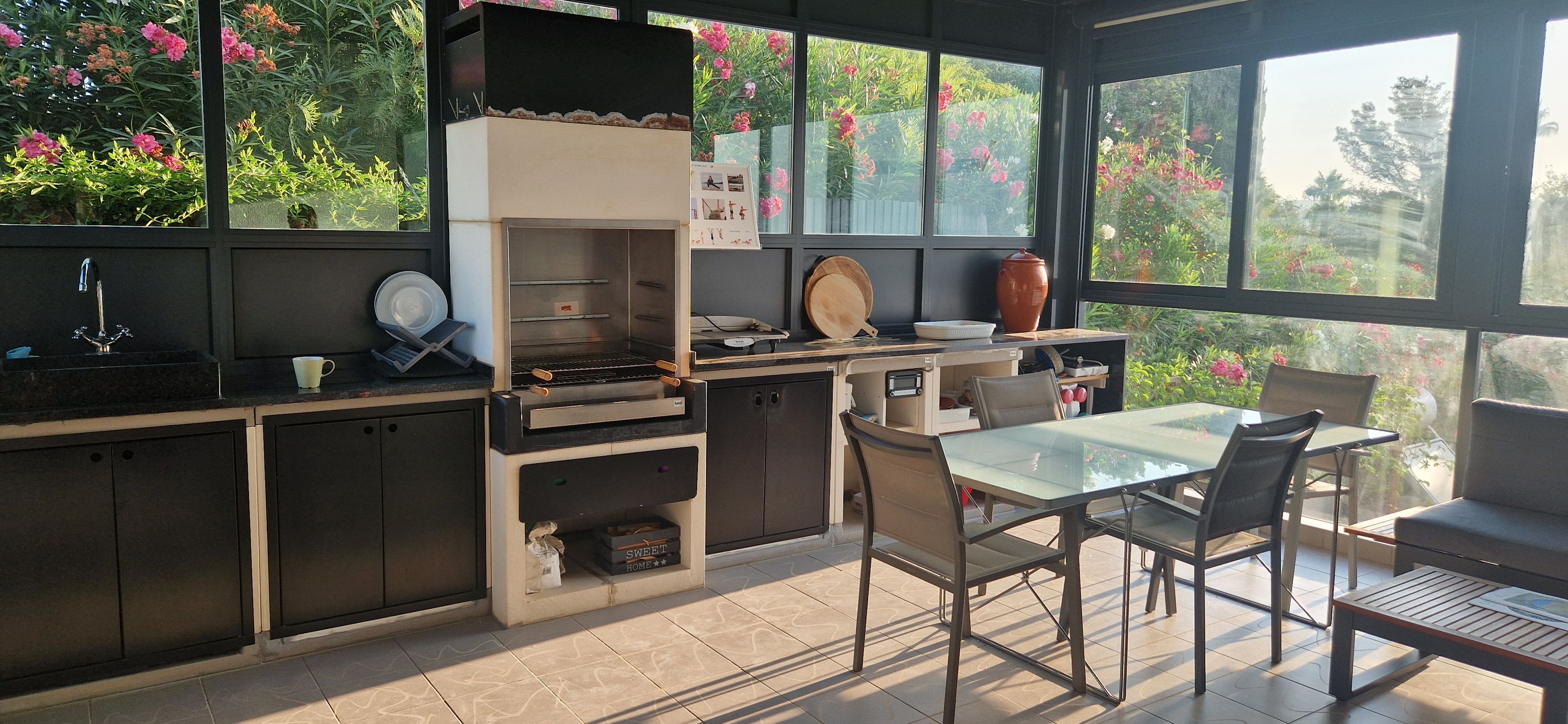 A kitchen with a built-in grill, a dining table with chairs, and a view of a garden through large windows.
