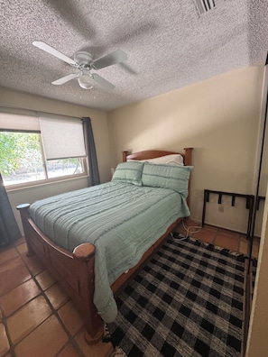 Een slaapkamer met een bed, een plafondventilator, een raam met zonwering en een geruit tapijt.